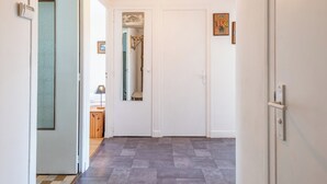 A hallway with a checkered floor, a white door, and a mirror reflecting a room with a bed and a lamp.