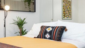 Een net opgemaakt bed met een gekleurd kussen, een bedlampje, een pot met plant en een kleurrijk kunstwerk aan de muur.