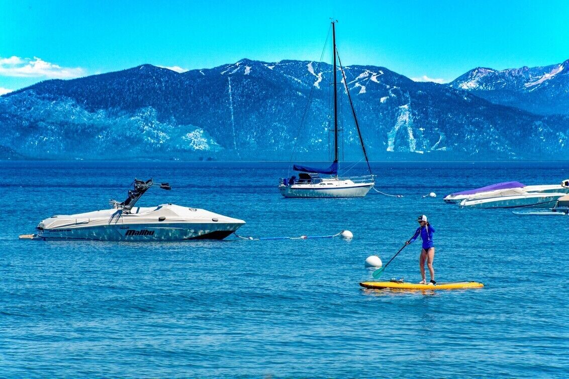 A person paddleboarding on a calm lake with boats and mountains in the background.