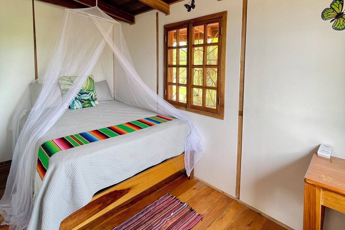 A room with a bed under a wooden ceiling, a window with wooden frames, and a mosquito net.