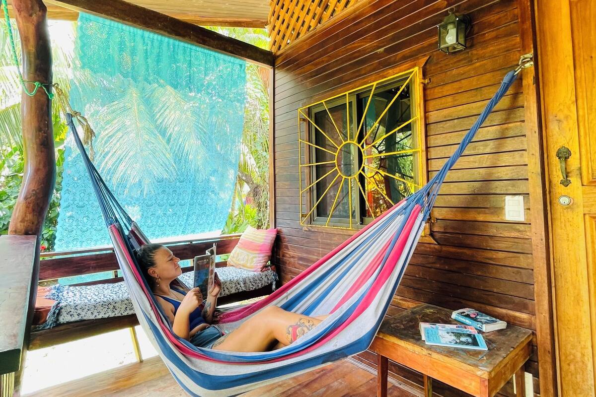 A hammock in a wooden room with a wooden ceiling and a window with a yellow frame.