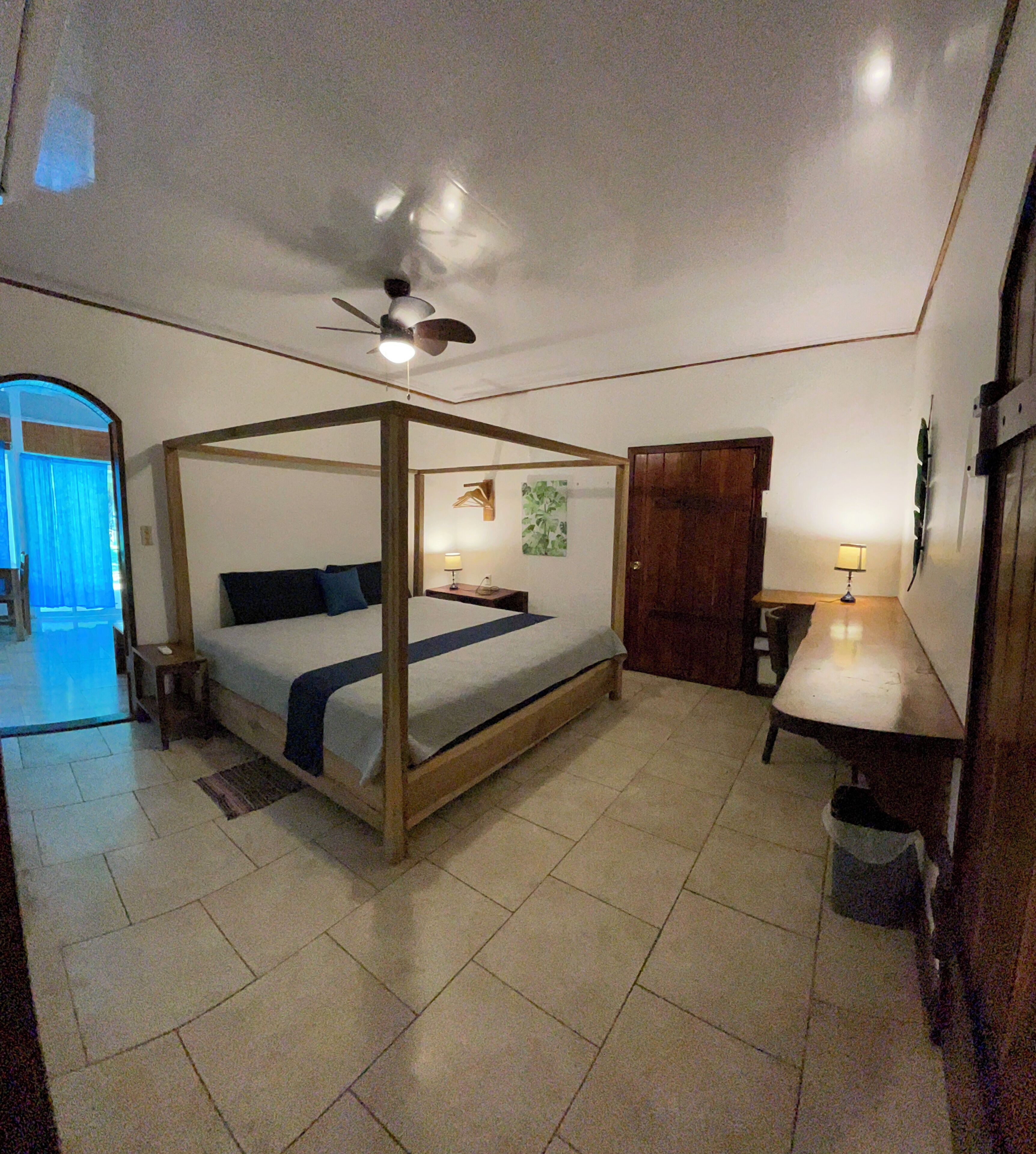 Un dormitorio con cama con dosel, ventilador de techo, mesita de noche con lámpara y puerta que da a otra habitación.