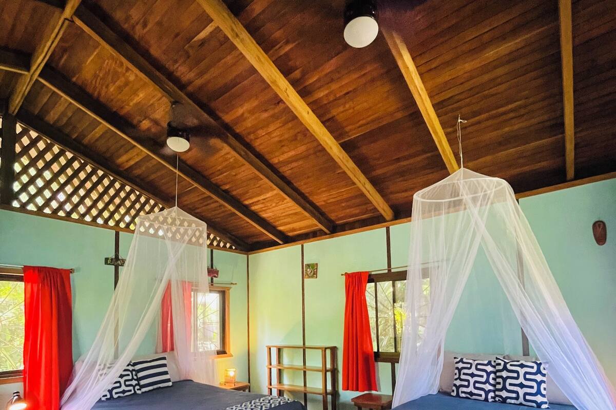 A room with two beds, wooden flooring, a wooden ceiling, and a window with red curtains.