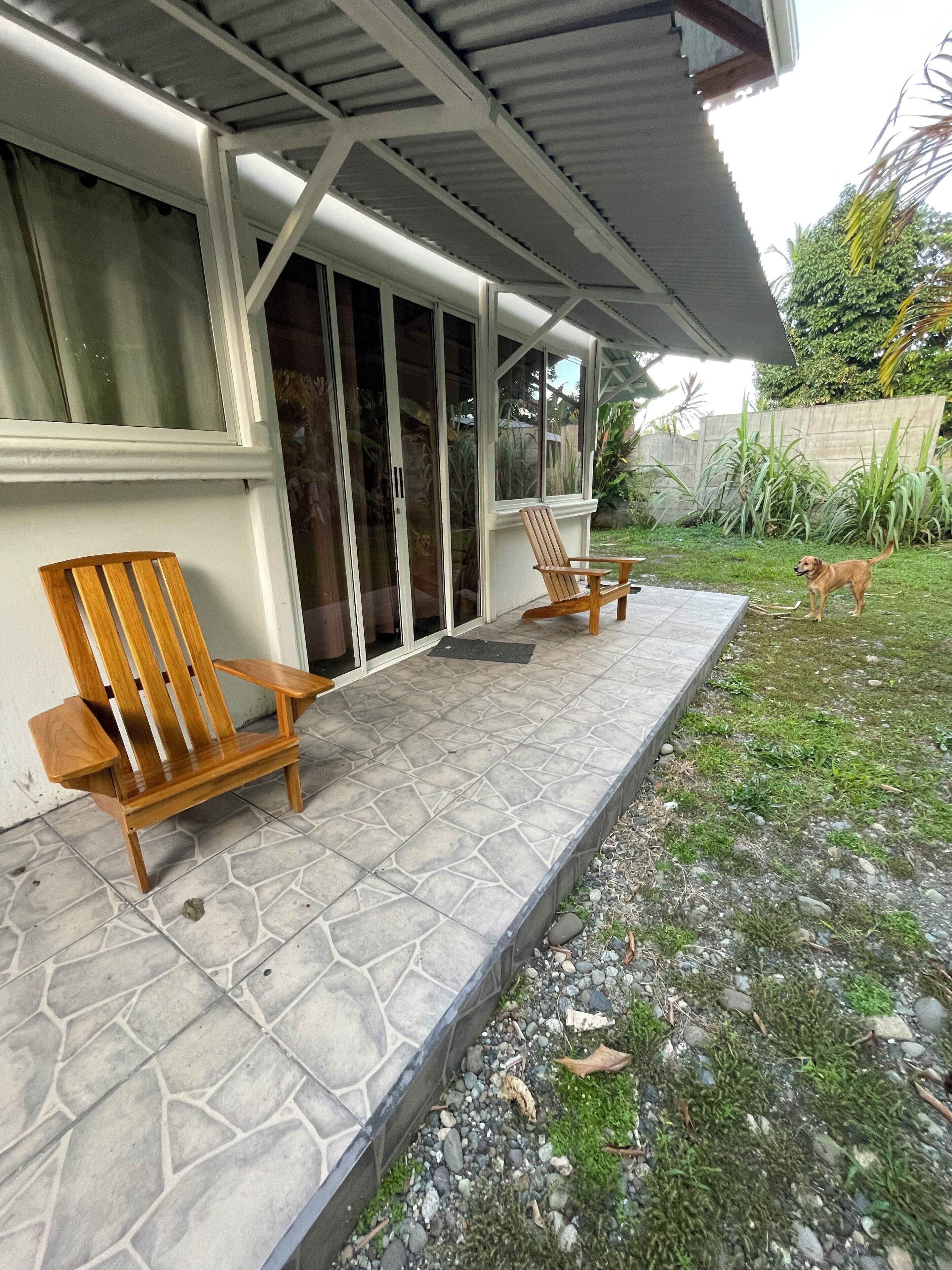 A covered patio with two wooden chairs, a tiled floor, and a grassy area with a dog.