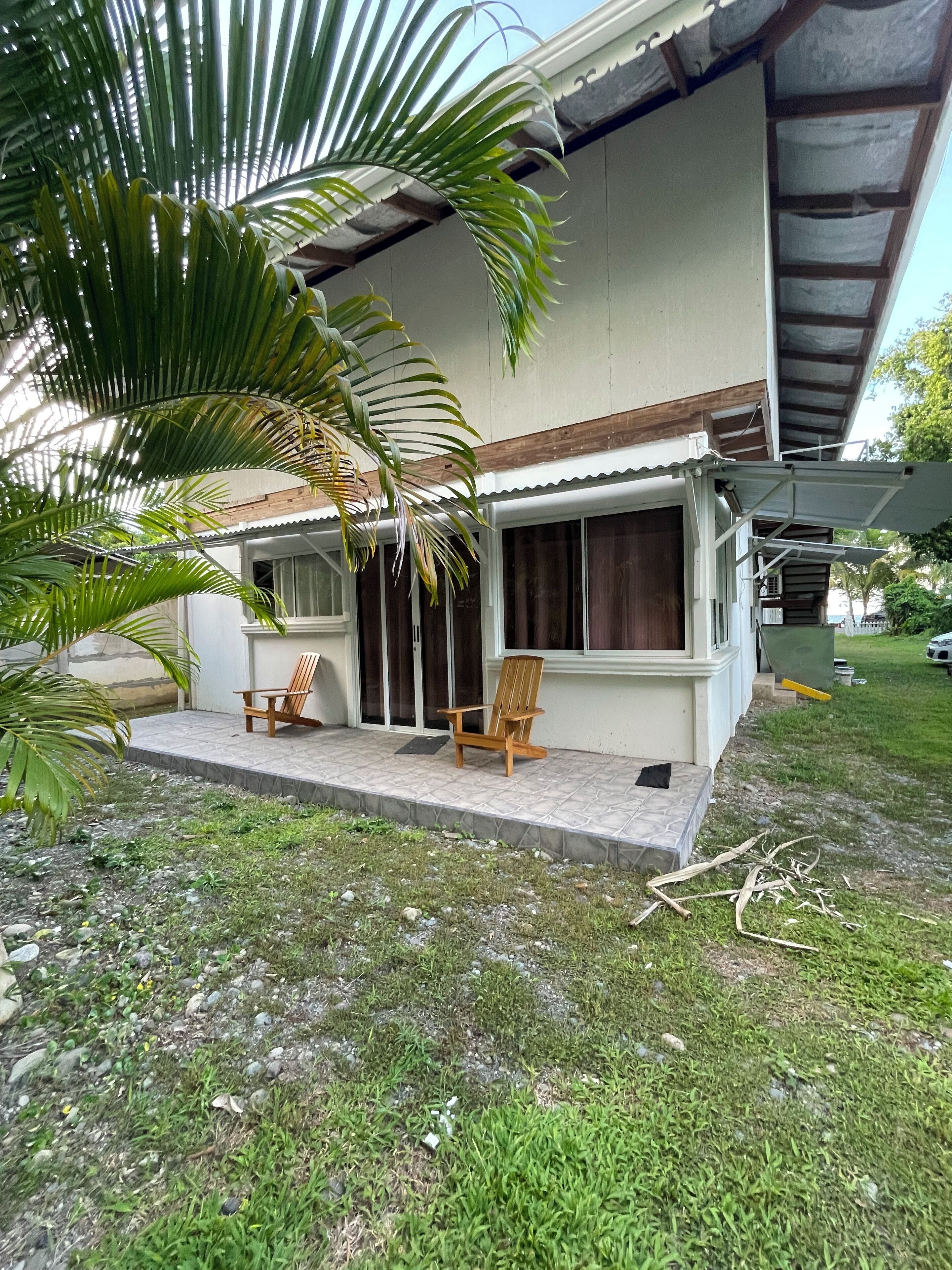 Ein Haus mit einer Veranda, zwei Holzstühlen und einer Palme.