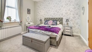 Quarto com cama, mesas de cabeceira, cômoda, janela com cortinas, radiador e um objeto decorativo na parede.