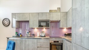 Uma cozinha moderna com armários cinzas, forno embutido e micro-ondas. Há um relógio na parede e uma cadeira azul em primeiro plano.