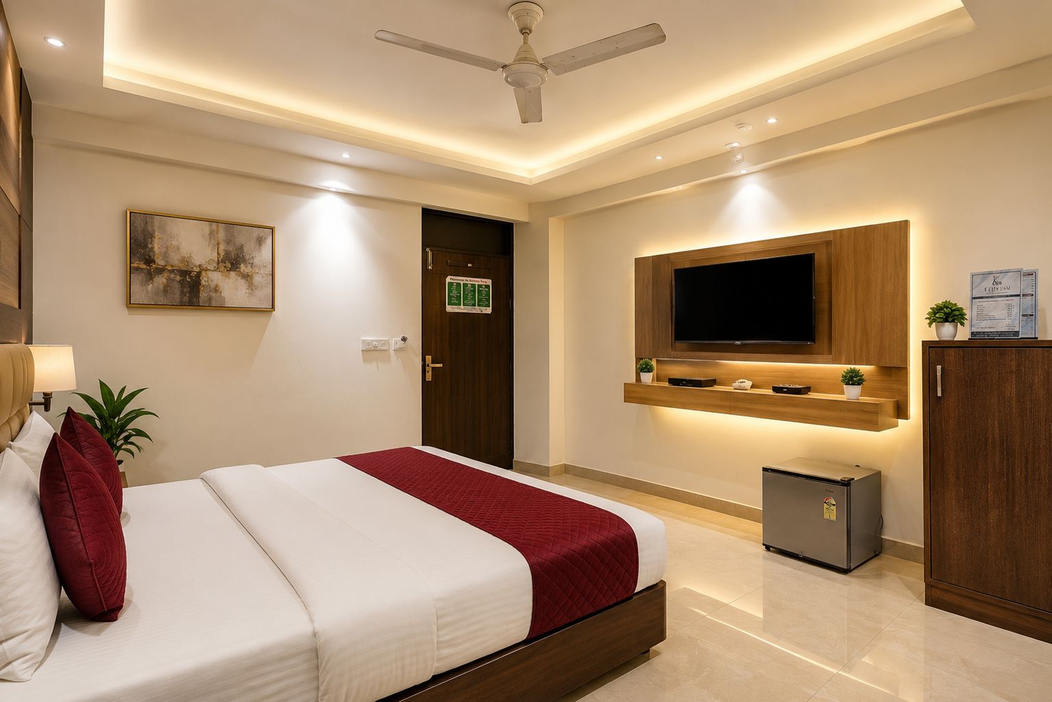 A hotel room with a bed, a TV mounted on the wall, a ceiling fan, and a small refrigerator.