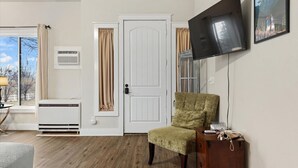 A room with a white door, a mounted TV, a chair, a wooden dresser, and a window with curtains.