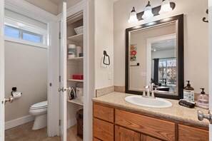 A bathroom with a toilet, open storage cabinets, a sink with a mirror, and toiletries.