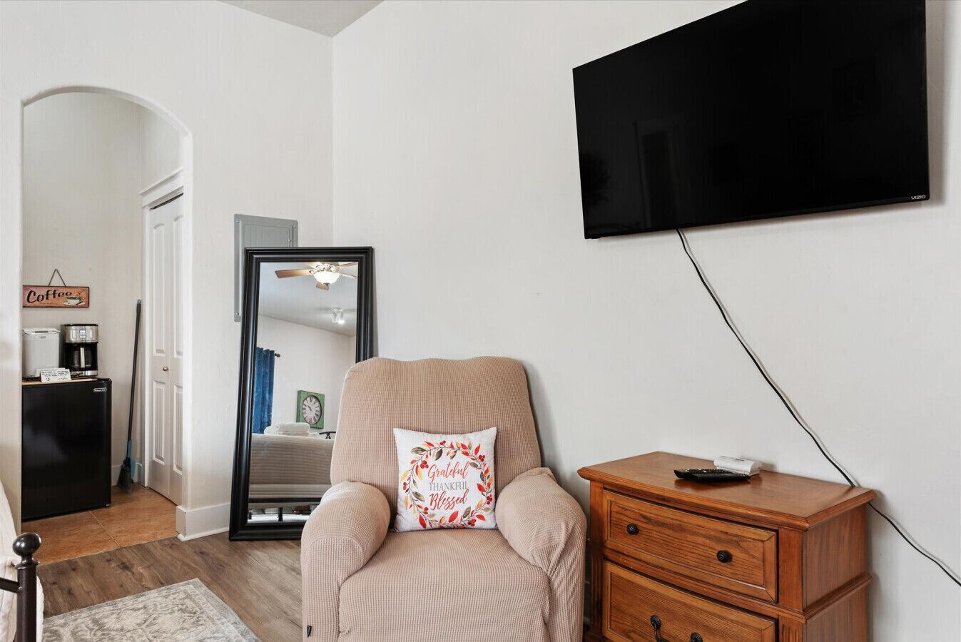 A living room with a beige armchair, a wooden side table, a flat-screen TV mounted on the wall, a coffee maker, and a clock on the wall.