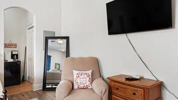 A living room with a beige armchair, a wooden side table, a flat-screen TV mounted on the wall, a coffee maker, and a clock on the wall.