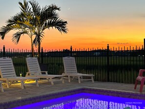 A pool area with lounge chairs and a palm tree, surrounded by a fence and a sunset view.