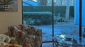 Una sala de estar con un sofá estampado, una mesa de centro de vidrio y vista a un jardín por un gran ventanal corredizo.