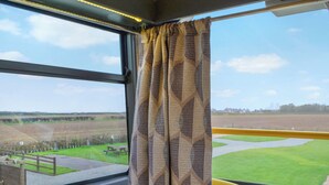 Une fenêtre ornée de rideaux à motifs, un couvre-lit jaune à motifs et une vue sur un champ verdoyant et une table de pique-nique.