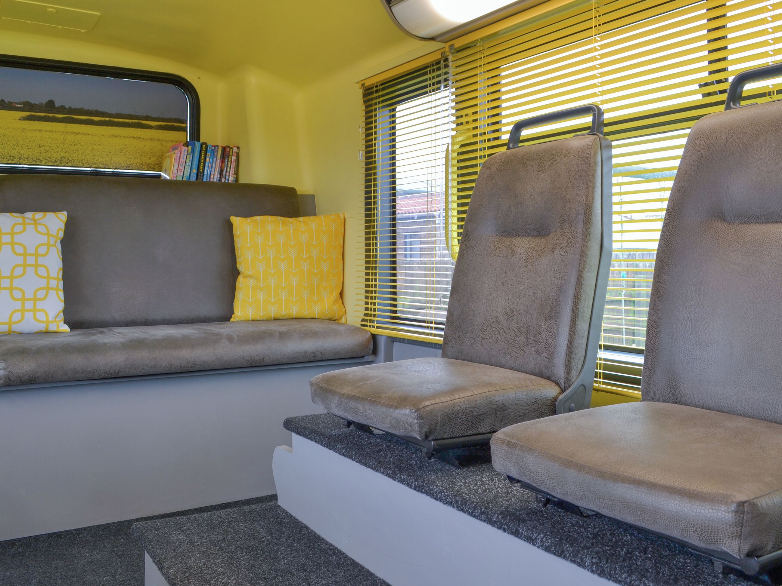 L'intérieur d'un wagon de train, avec un espace pour s'asseoir, une étagère remplie de livres et une fenêtre équipée de stores.