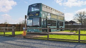 Un autobus à deux étages arborant la marque « Daisy Decker », garé à un arrêt de bus, avec une clôture en bois et une table de pique-nique en arrière-plan.