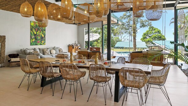 A dining area with wicker chairs and a large table, surrounded by large windows with a view of greenery and a pool.