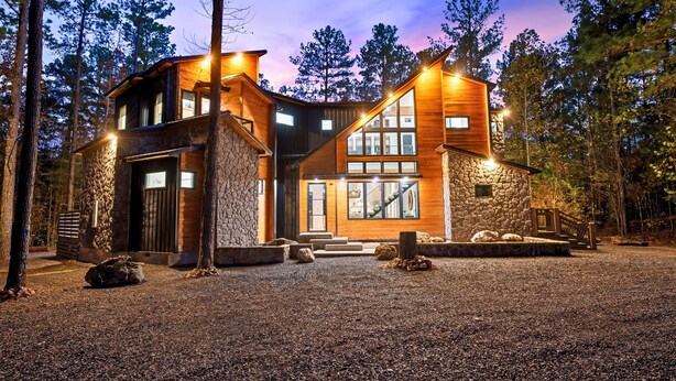 A modern log cabin with a stone foundation, large windows, and a gravel driveway surrounded by trees. - Luxury Creekside Lodge for Group Gatherings (Broken Bow)