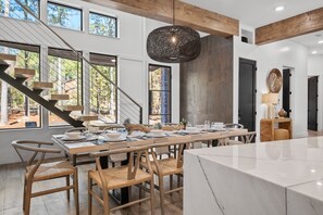 Un espace repas moderne avec une longue table dressée pour six personnes, des chaises en bois, un escalier et de grandes fenêtres donnant sur la forêt.