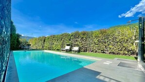 Ein Schwimmbecken mit klarem, blauem Wasser, umgeben von einer gepflegten Hecke und einigen Liegestühlen.