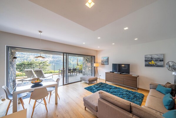 A modern living room with a dining area, a sofa, a TV, and a view of the outdoors. - BELLAVISTA LAGLIO LAGOON B by Italian Apartments (LAGLIO)