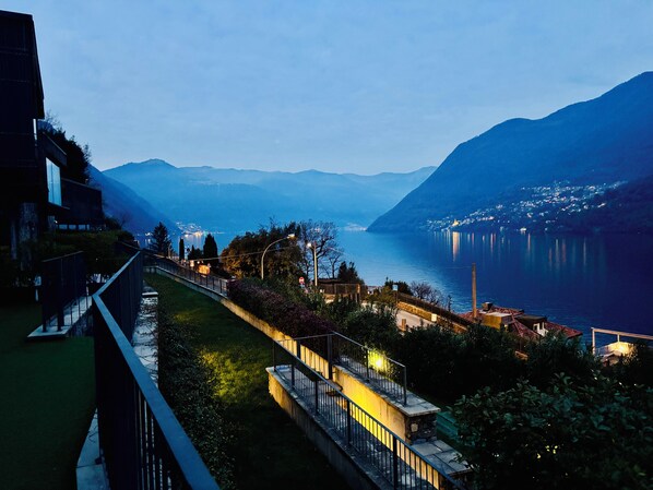 A lakeside view with mountains in the background, a pathway, and a building with a balcony overlooking the water. - BELLAVISTA LAGLIO LAGOON B by Italian Apartments (LAGLIO)