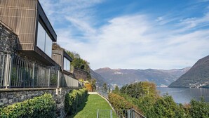 Ein modernes Gebäude mit einer Steinaußenwand, einem grünen Weg und im Hintergrund eine Seen- und Berglanschaft.