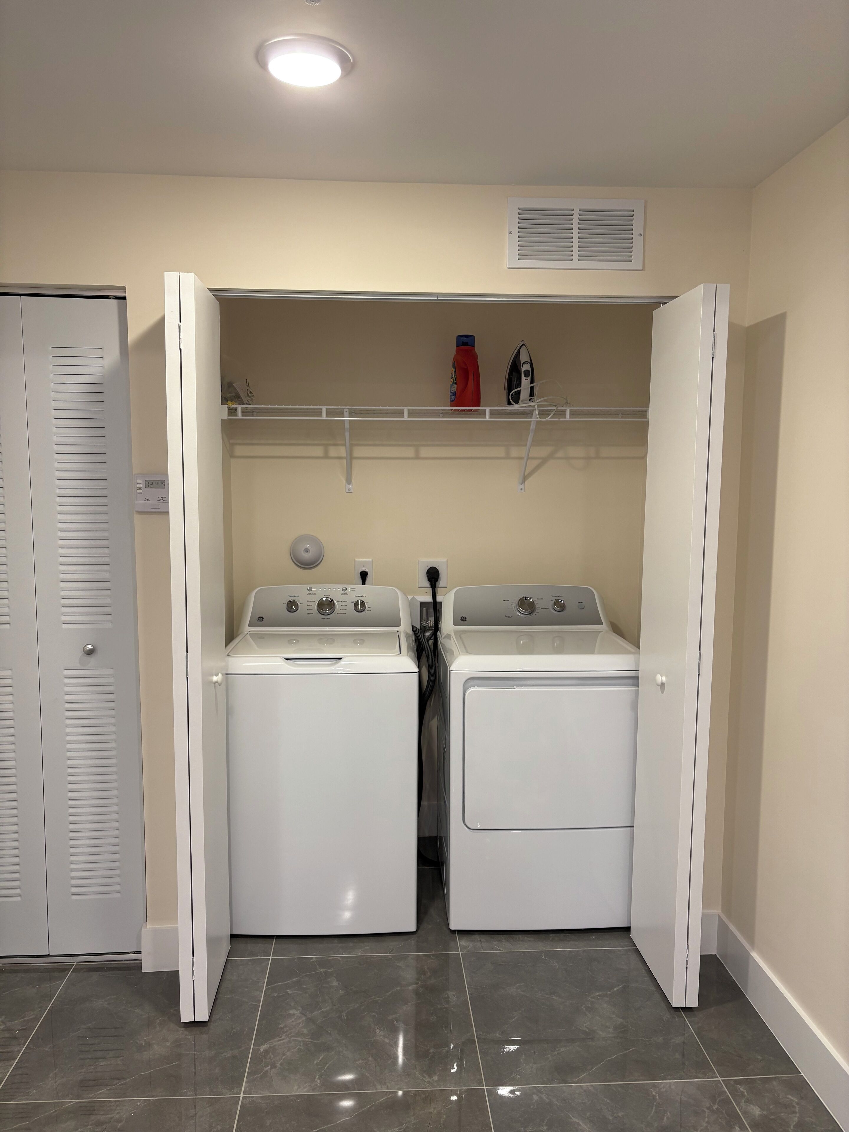 A laundry room with two white front-loading washers and dryers, a shelf above with a red bottle and an iron, and a vent on the wall.