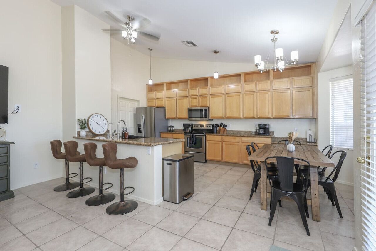 A modern kitchen with wooden cabinets, stainless steel appliances, a central island with bar stools, and a dining area with black chairs.