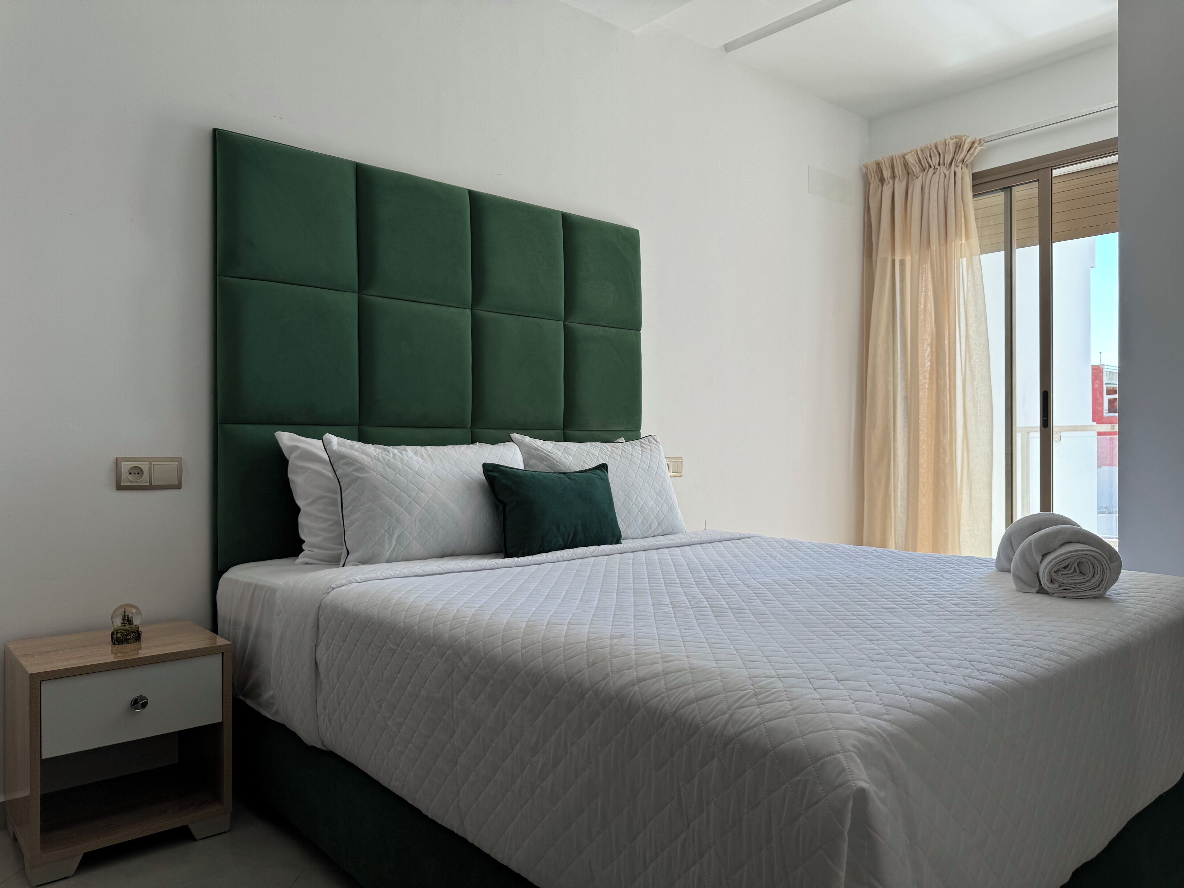 Een net opgemaakt bed met een groen gestoffeerd hoofdeinde, witte beddengoed en een klein nachtkastje met een lamp.