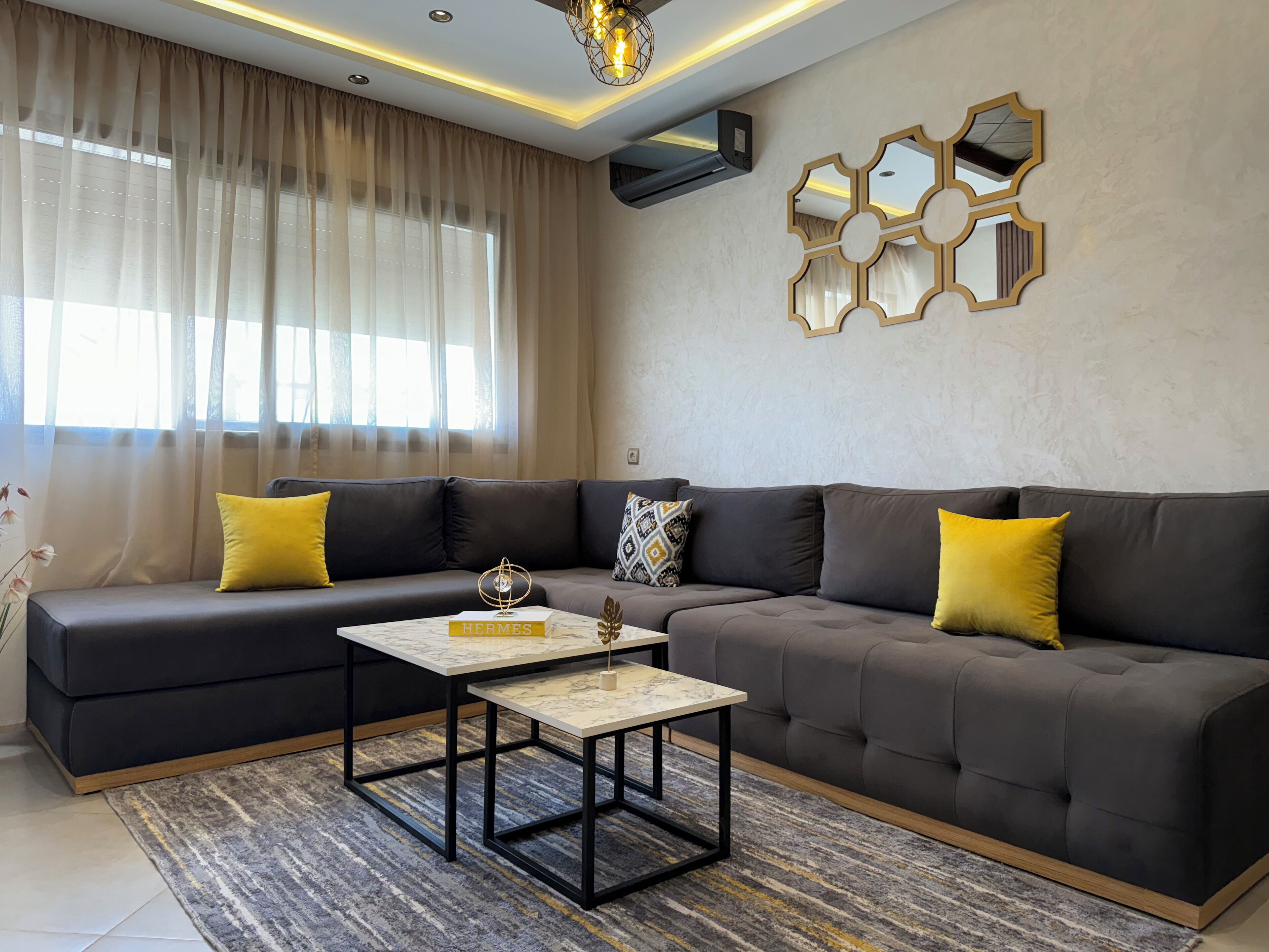 Un salon moderne avec un canapé gris foncé, des coussins jaunes, une table basse en marbre et un tapis à motifs.