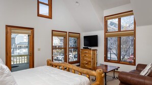 Ein Schlafzimmer mit Bett, Holzkommode, Fernseher und großen Fenstern mit Blick ins Freie.