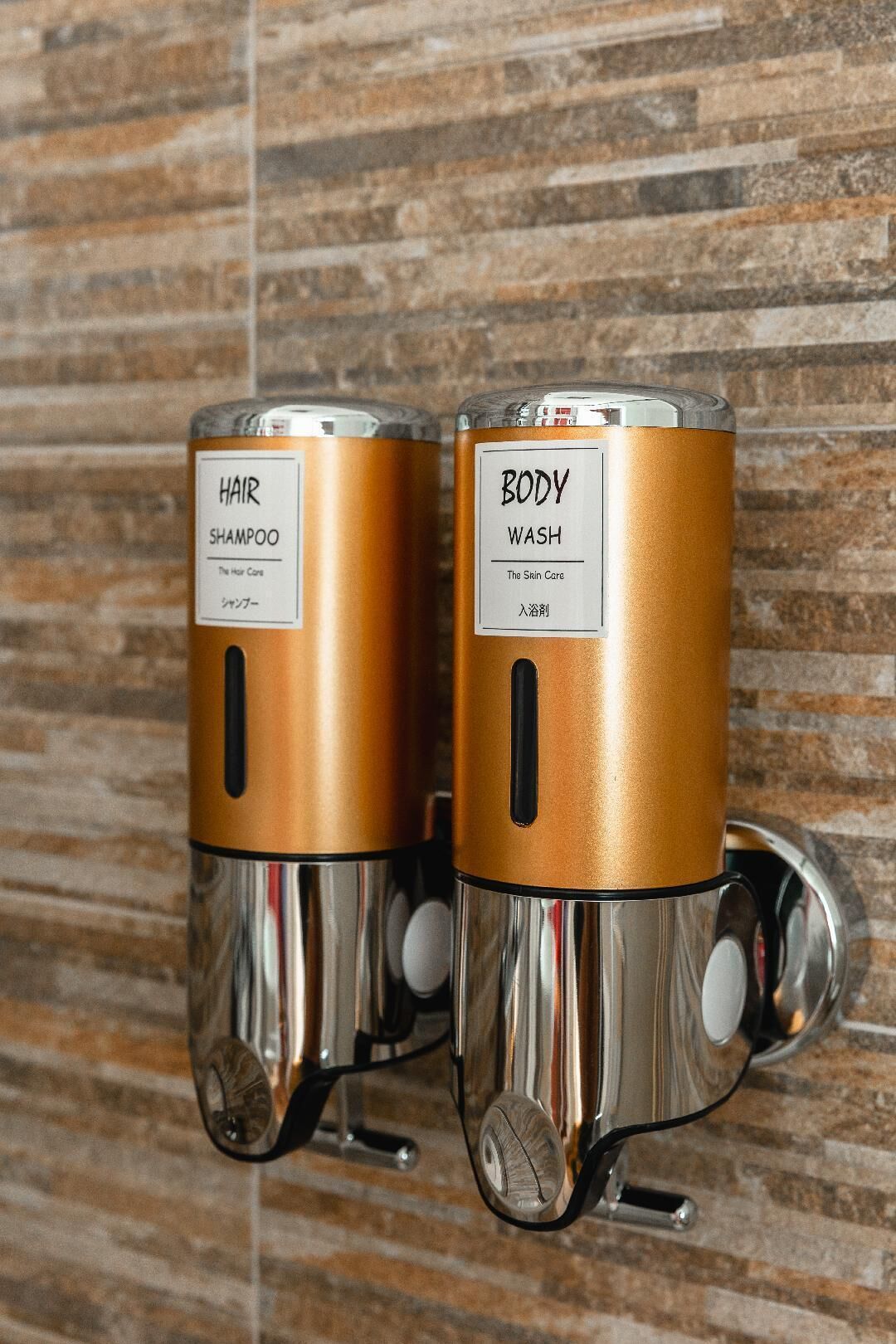 Zwei an der Wand montierte, metallische, zylinderförmige Spender mit den Aufschriften 'HAIR SHAMPOO' und 'BODY WASH' an einer strukturierten Wand.