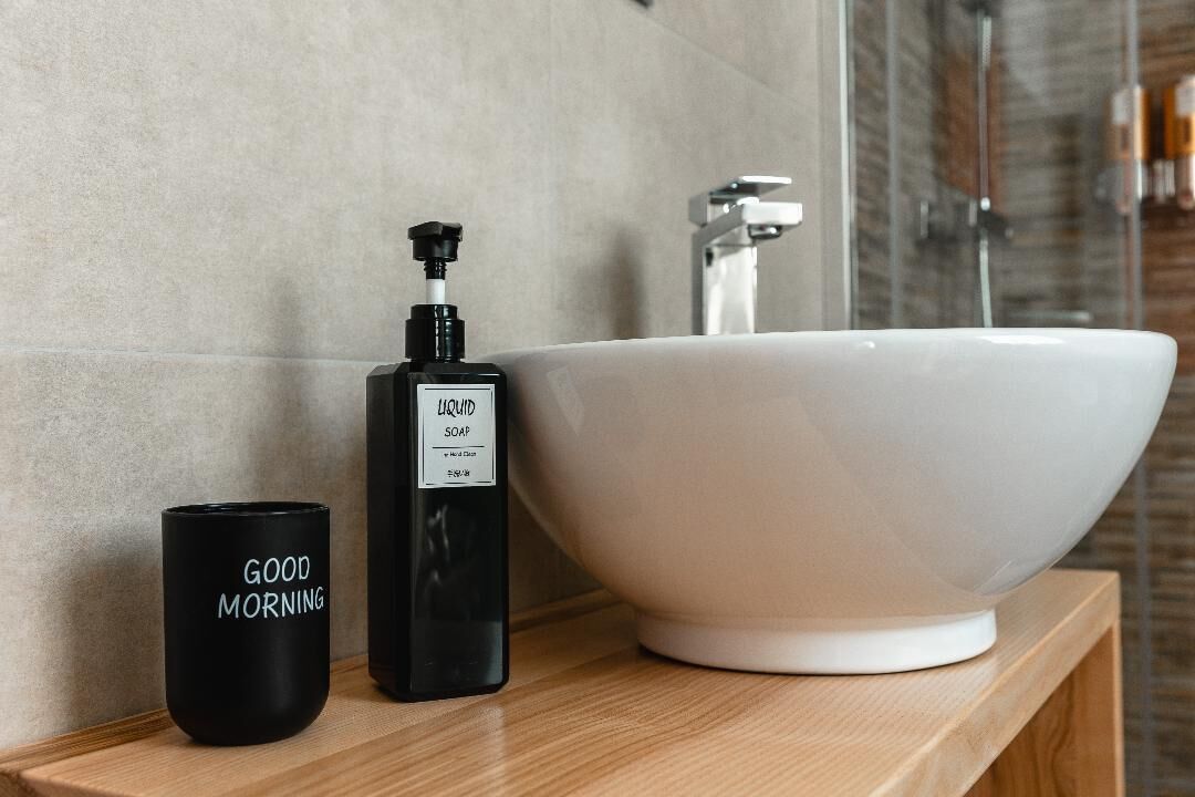 Une salle de bain avec un lavabo blanc, un distributeur de savon noir et une tasse noire sur laquelle est écrit « BONJOUR ».