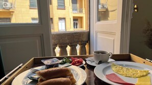 A breakfast tray with croissants, ham, cheese, and an omelette, along with a cup of coffee and yogurt, on a wooden table by a window with a view of a building. - Charming Oasis in the Heart of Cairo – Authentic Hospitality Awaits! (Cairo Governorate)