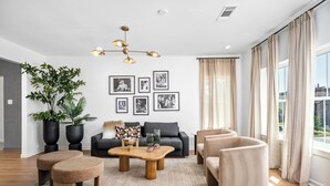 A modern living room with a grey sofa, a wooden coffee table, and a beige armchair. There are large potted plants, a chandelier, and framed pictures on the wall.
