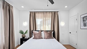 A bedroom with a large bed, beige curtains, a bedside table with a plant, and a wall-mounted light fixture.