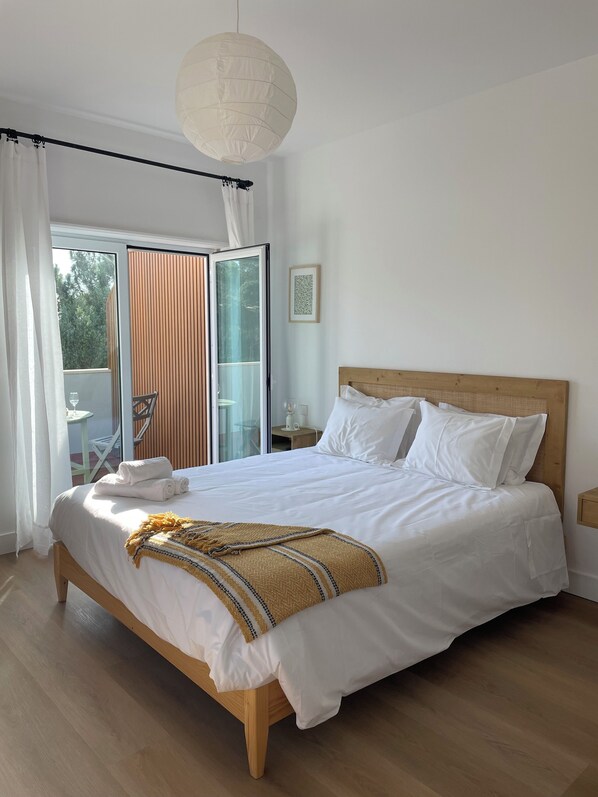 A bedroom with a large bed, a wooden headboard, a hanging light fixture, a sliding glass door to a balcony, and a framed picture on the wall.