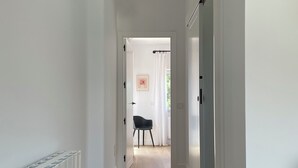 A narrow hallway with white walls and wooden flooring, leading to a room with a black chair and a framed picture on the wall.