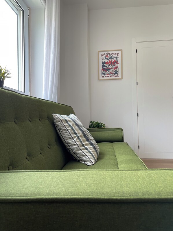 Um sofá verde com uma almofada listrada, um quadro na parede e um vaso de planta no parapeito da janela.