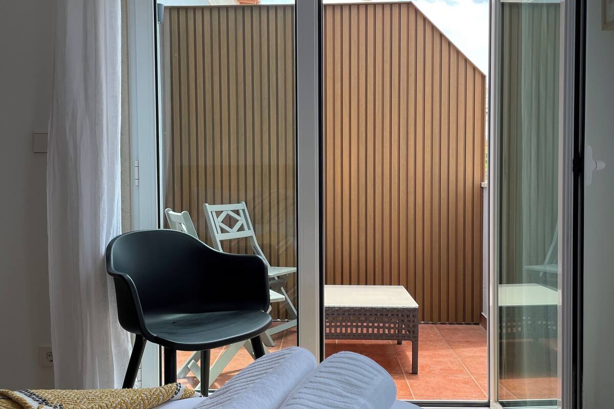 Ein Hotelzimmer mit Bett, Stuhl, kleinem Tisch und Blick auf einen Holenzaun durch eine Schiebetür.