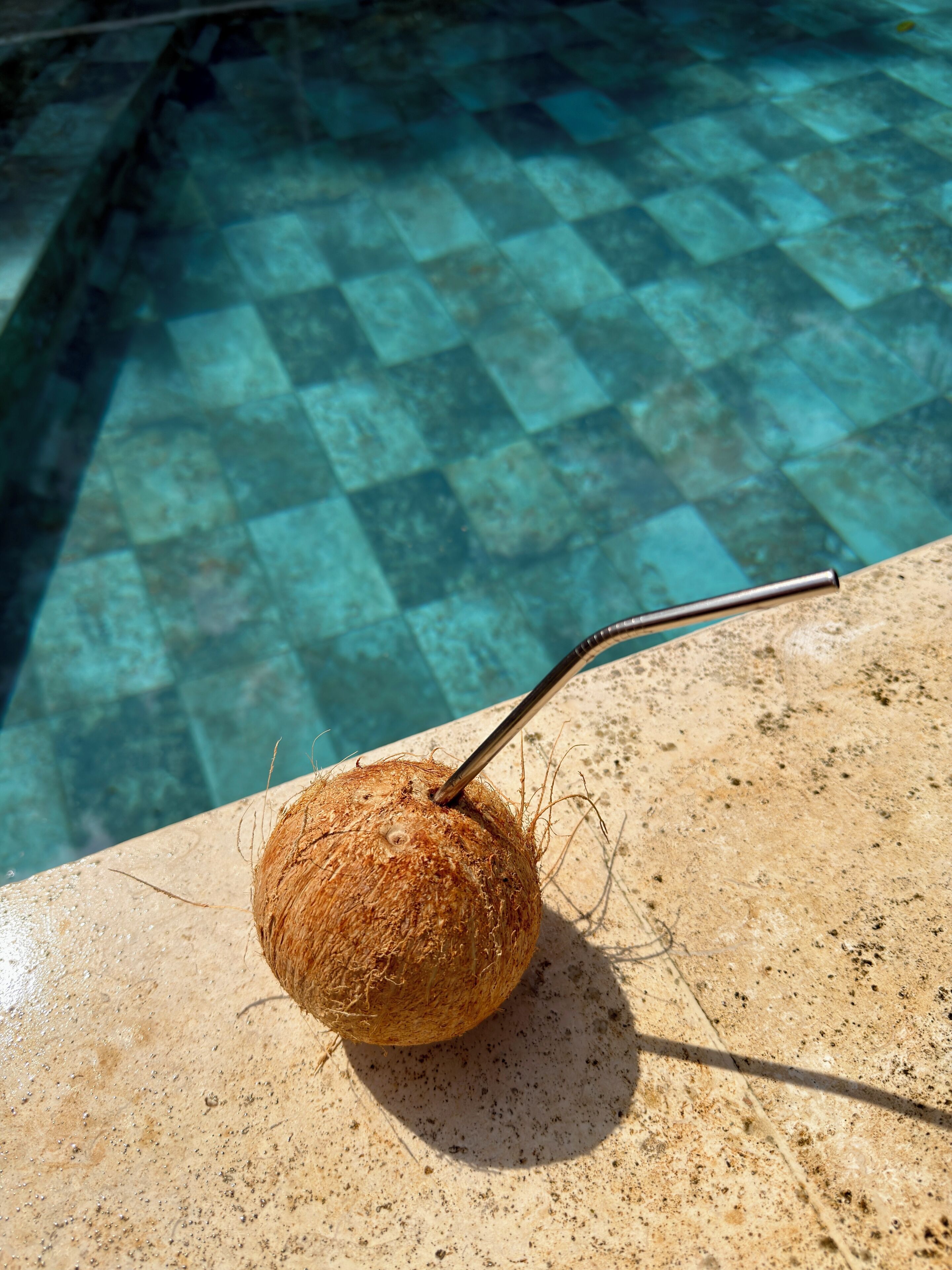 A coconut with a straw on a stone surface near a pool.