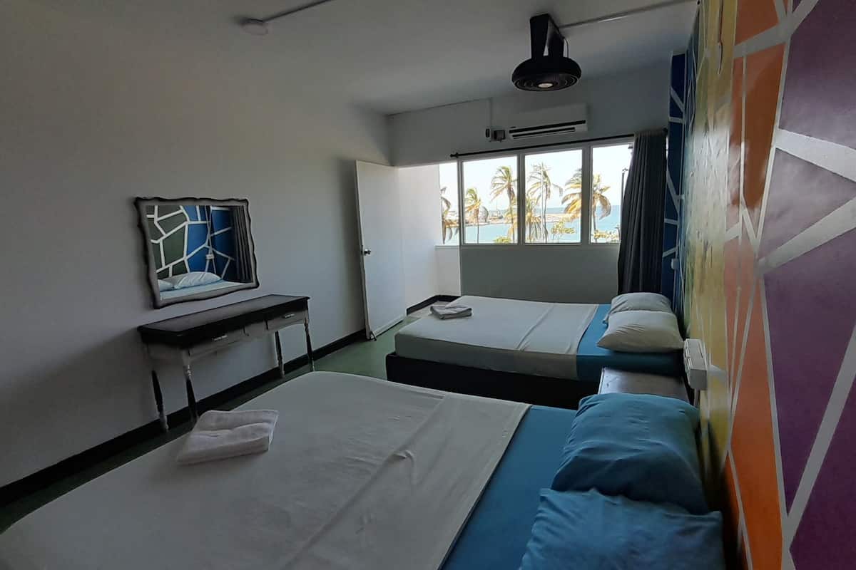 A hotel room with two beds, a desk, a mirror, and a window with a view of palm trees.