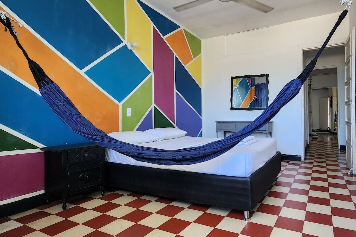 A room with a bed, a hammock, a colorful geometric wall, a checkered floor, and a hallway.