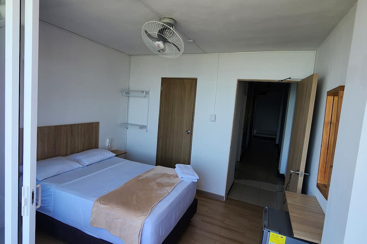 A hotel room with a bed, a bedside table, a fan, and a door leading to another room.