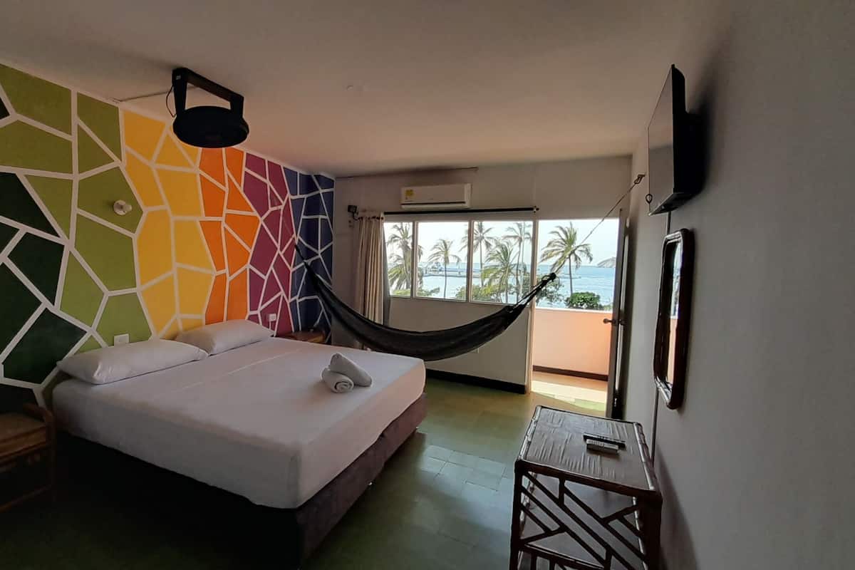 A hotel room with a bed, a hammock, a small table, and a window with a view of palm trees and the sea.