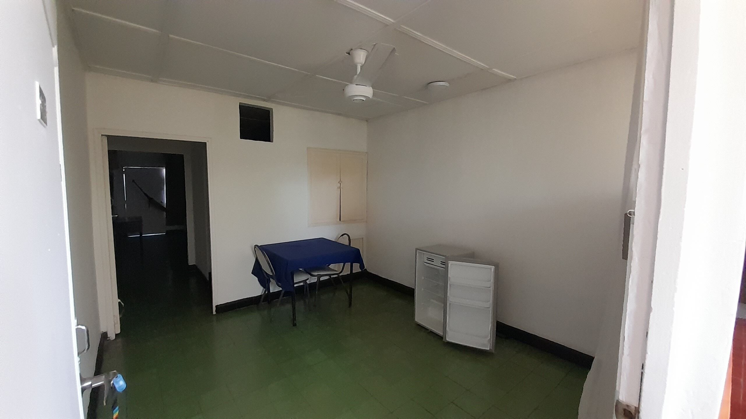 Una stanza con un letto singolo, una scrivania, una sedia, un piccolo armadietto bianco e una ventola a soffitto.