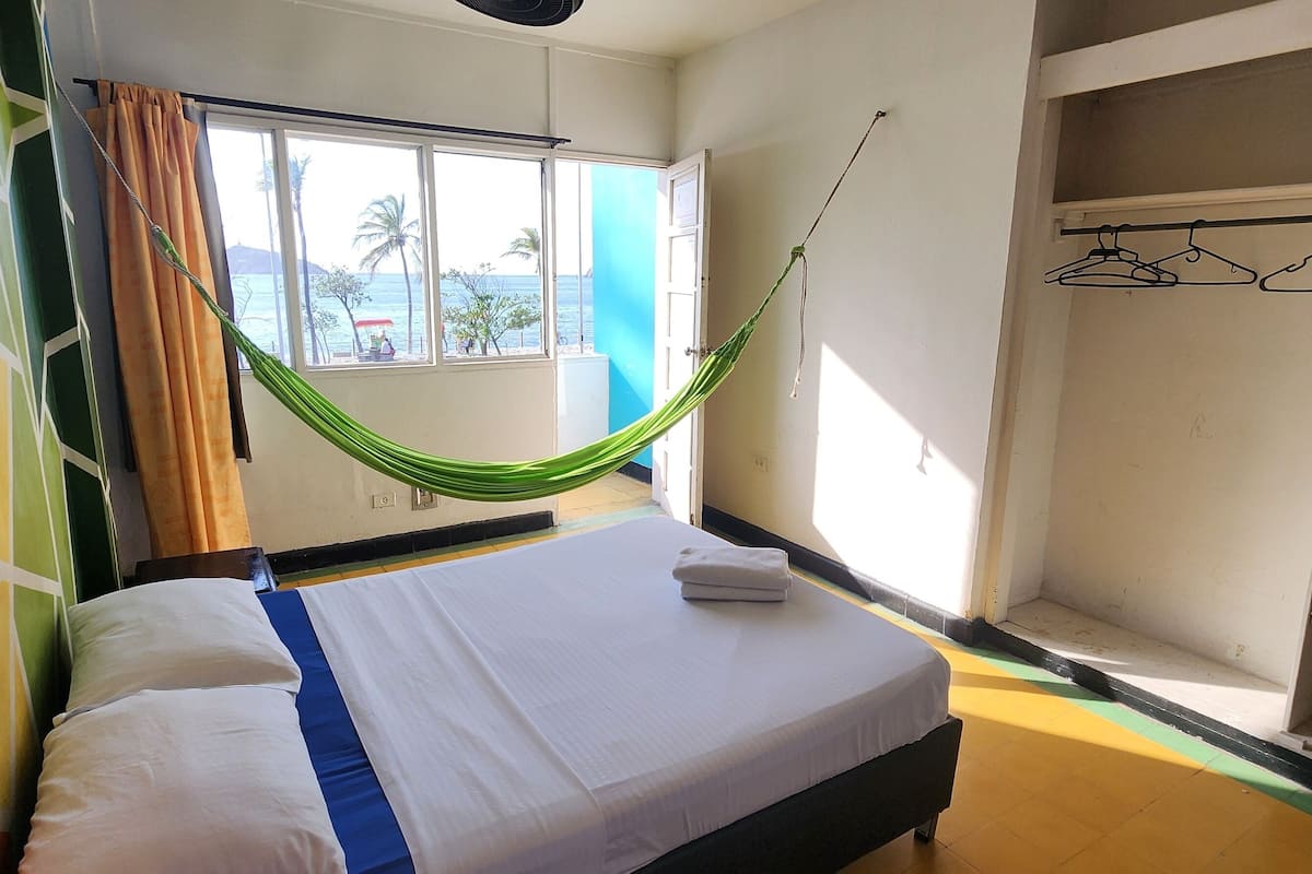 A room with a bed, a window with a view of the sea, a hammock, and a wardrobe.