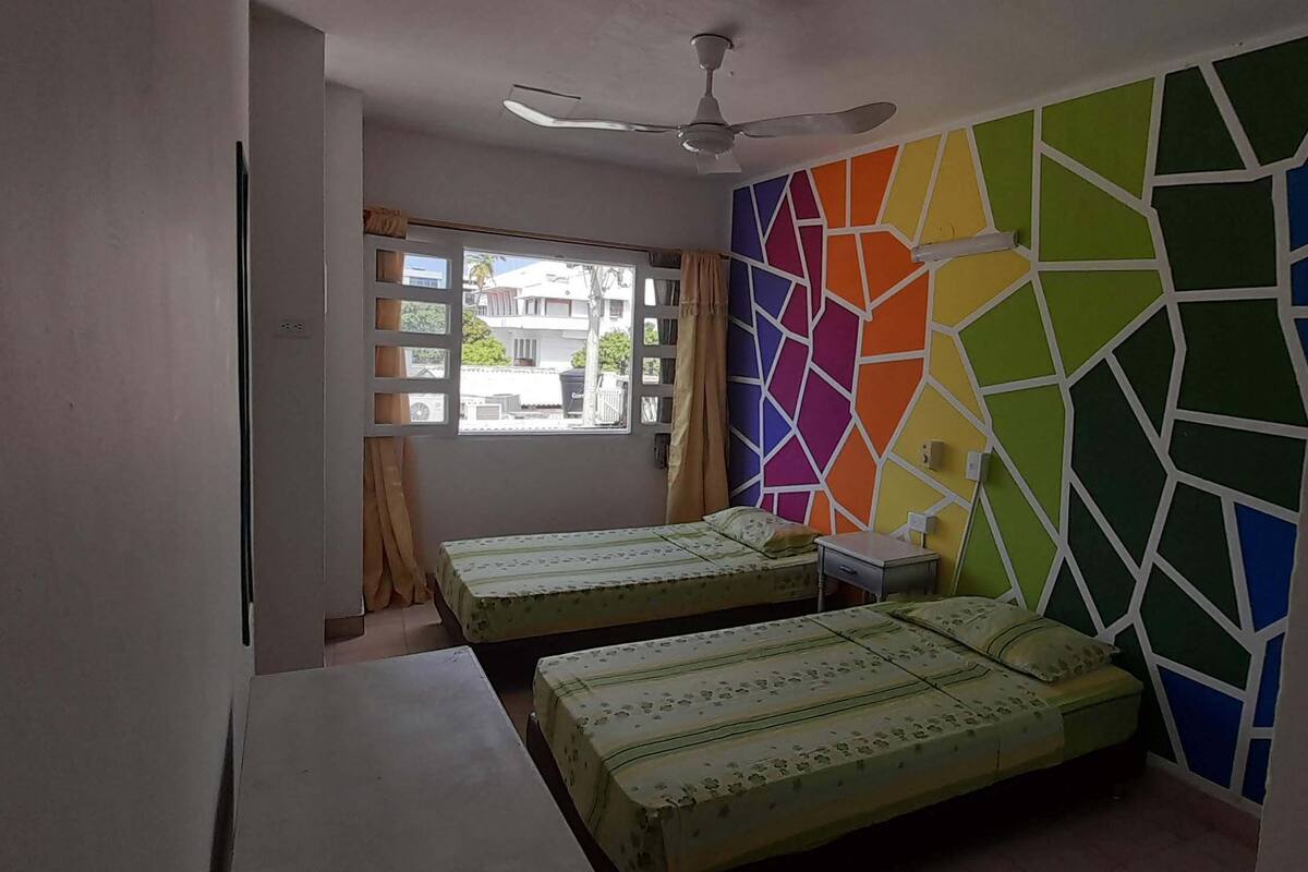 A room with two beds, a ceiling fan, a window with curtains, and a colorful wall.